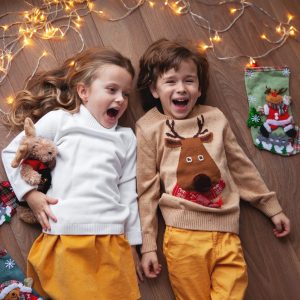 Children lying, laughing and have fun with Christmas decor, garland of light bulbs. Beautiful kids wearing sweater with deer. Overhead View, Close up portrait